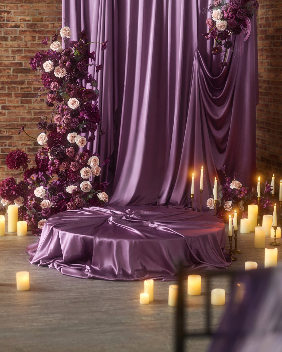 Purple satin draped platform with floral arrangements and glowing white pillar candles.
