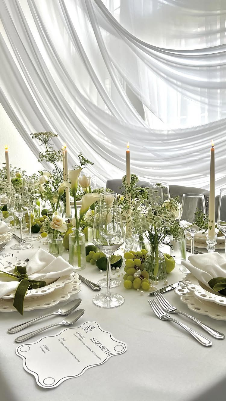 Wedding table setting with white linens, green floral arrangements, and white drapery background.