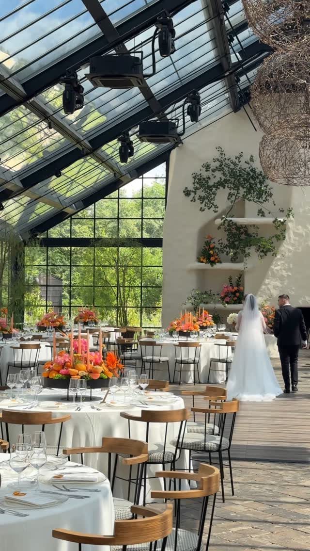 Wedding reception tables with bright orange floral centerpieces under a glass ceiling.