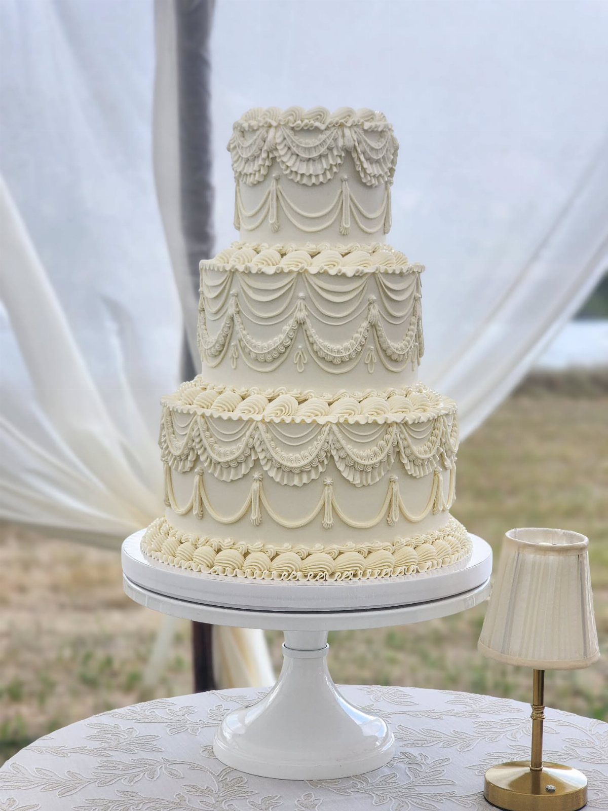Three-tiered white wedding cake with ornate frosting swags on a lace-covered table.