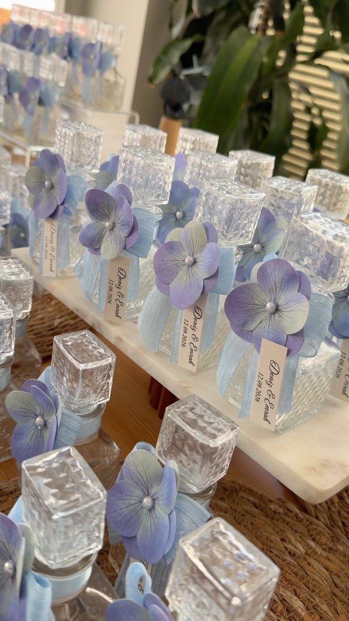 Small glass favor bottles with blue ribbons and purple hydrangea petals on marble.