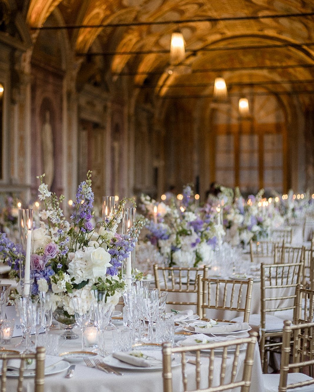 Floral centerpieces with purple and blue flowers on white wedding banquet tables.