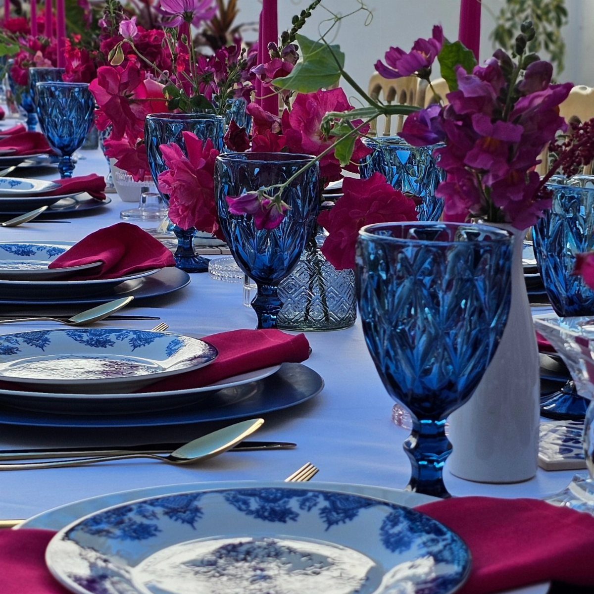 Blue patterned plates and glassware with magenta napkins and purple flowers on a table.