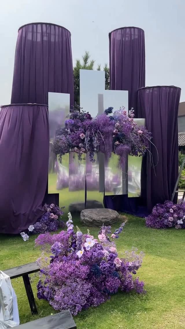 Purple floral arrangements and fabric drapes surrounding mirrored panels on a green lawn.