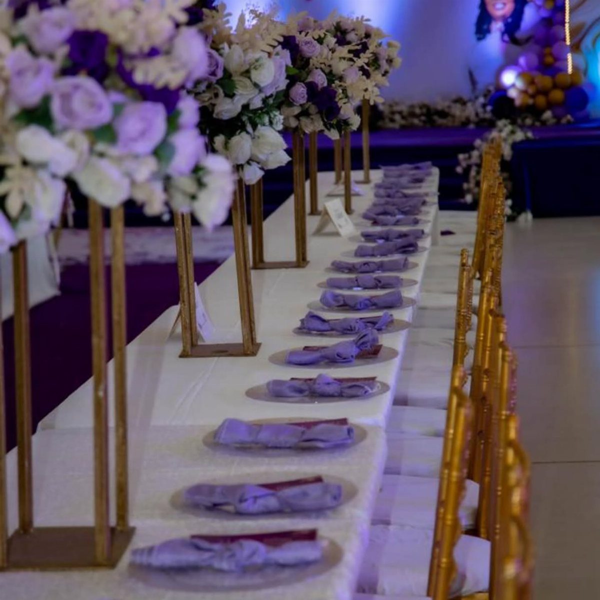 Purple and white floral centerpieces on gold stands atop a white wedding table.