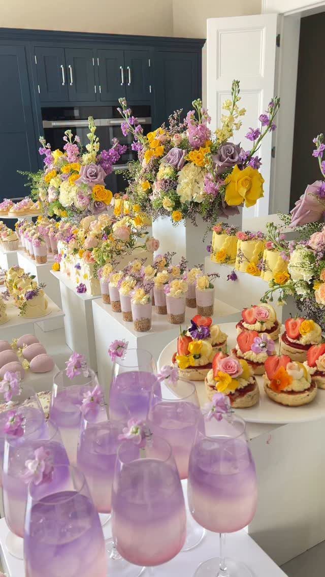 Dessert table with purple and yellow flowers, purple drinks, and assorted pastries.