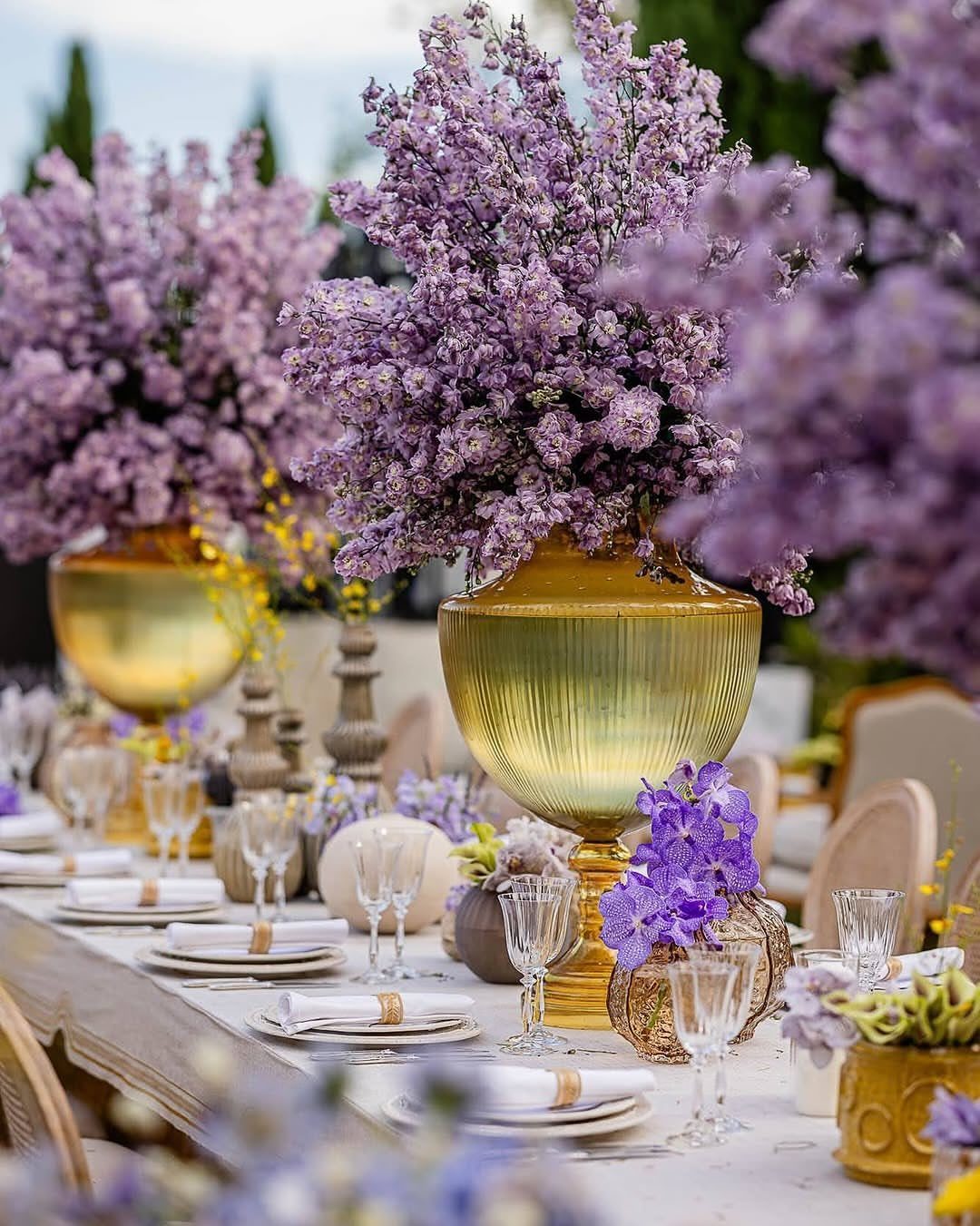 Purple floral arrangements in gold vases on a white wedding reception table.