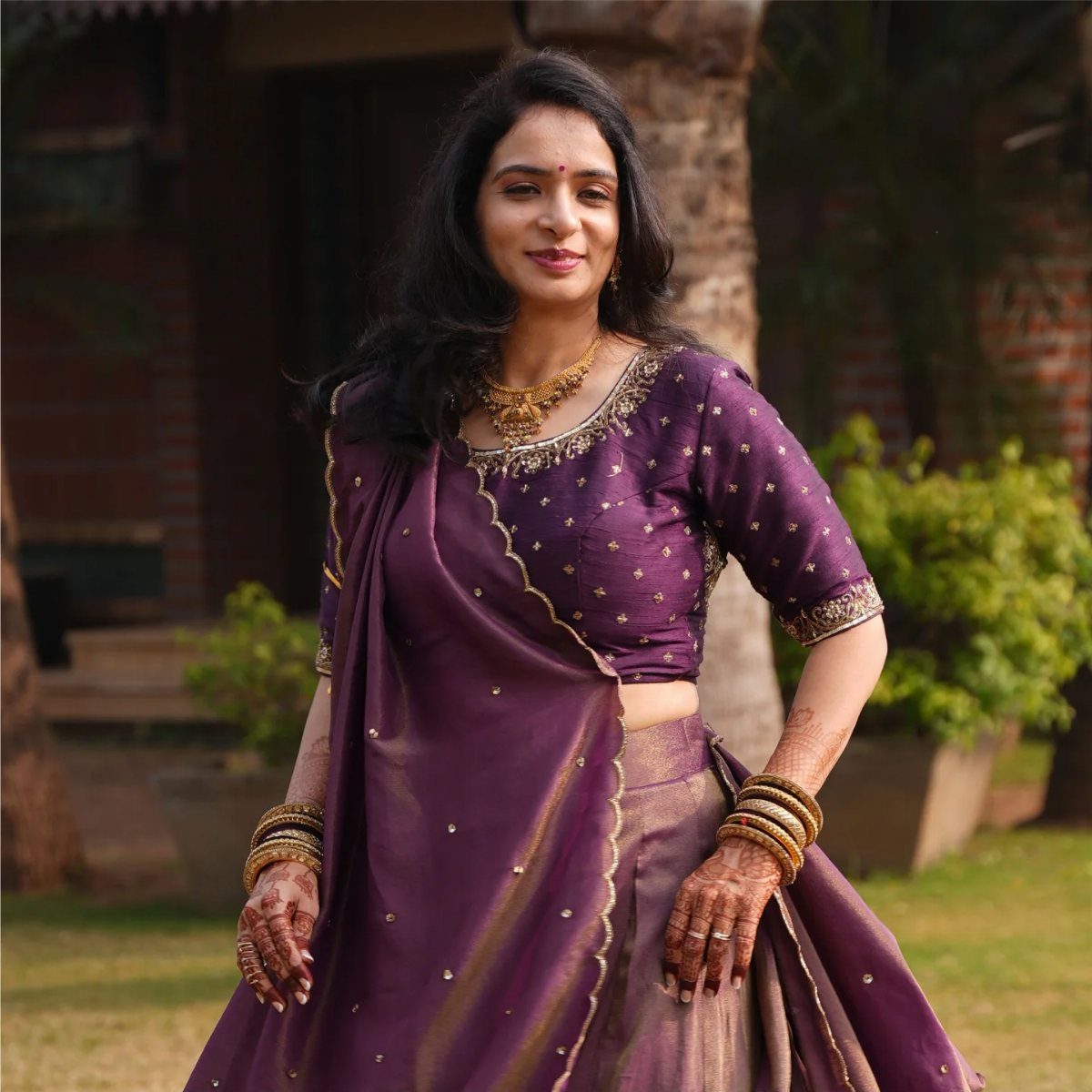 Woman in purple and gold embroidered Indian lehenga posing in a green garden