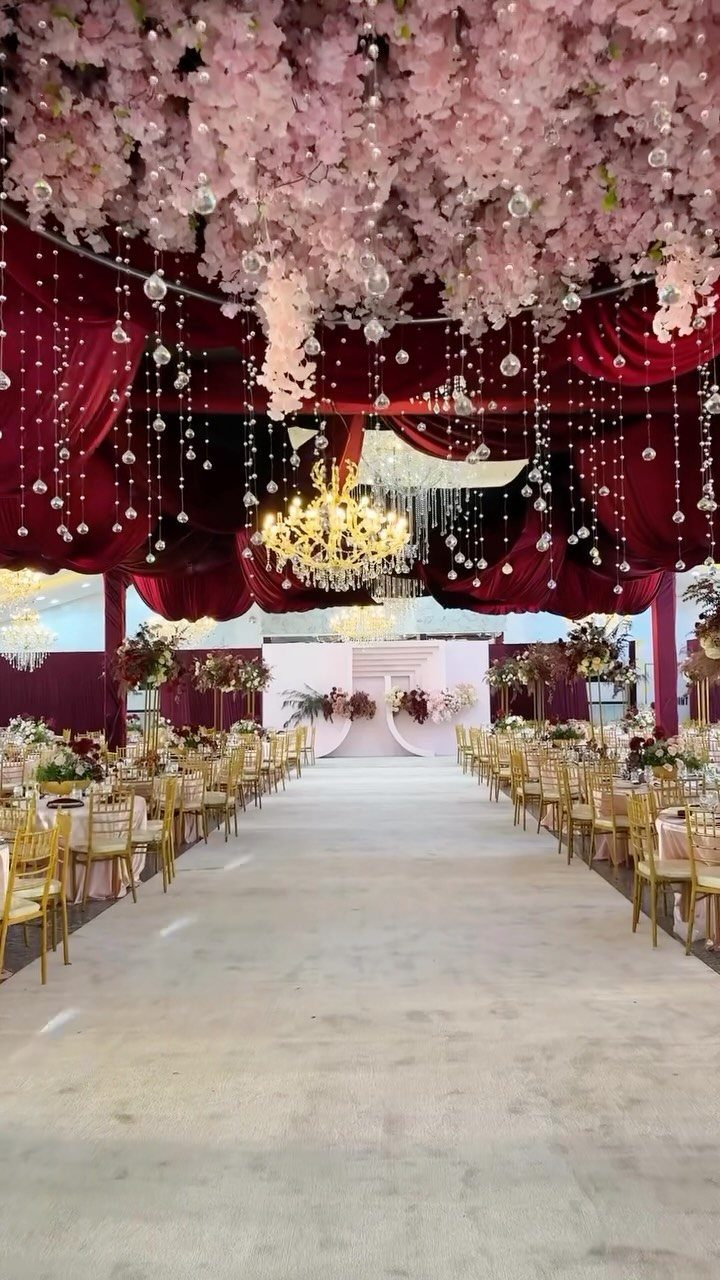 Wedding reception hall with burgundy velvet curtains, pink floral ceiling, and gold chairs.