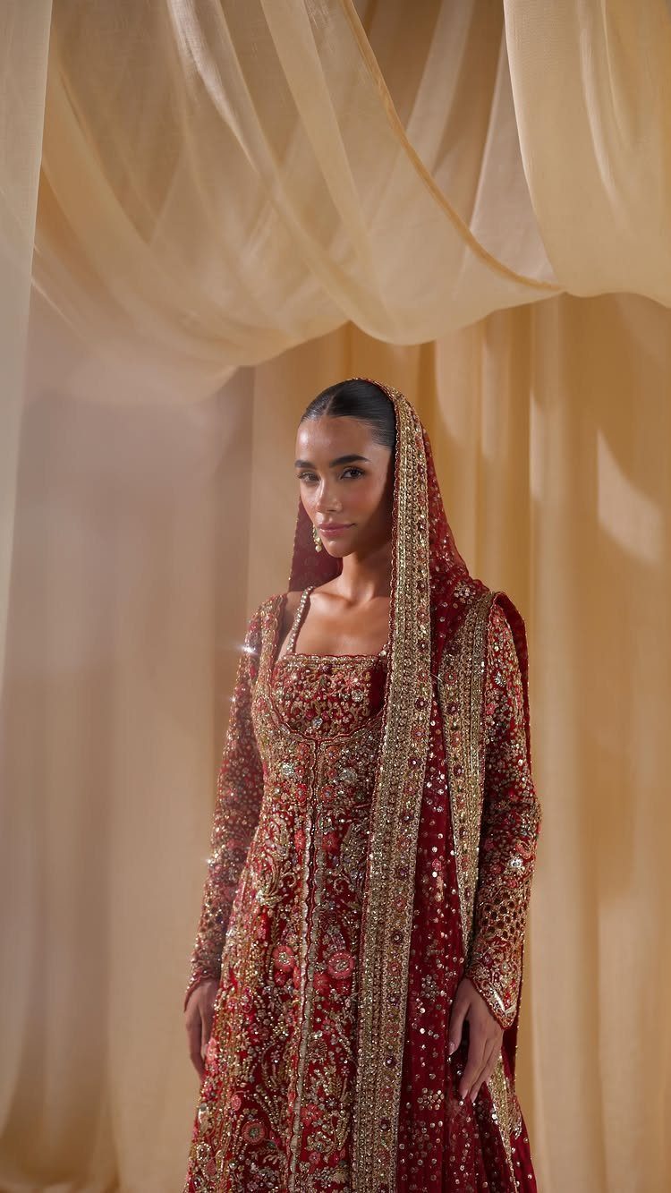Woman in an embroidered red and gold bridal gown against cream fabric drapes.