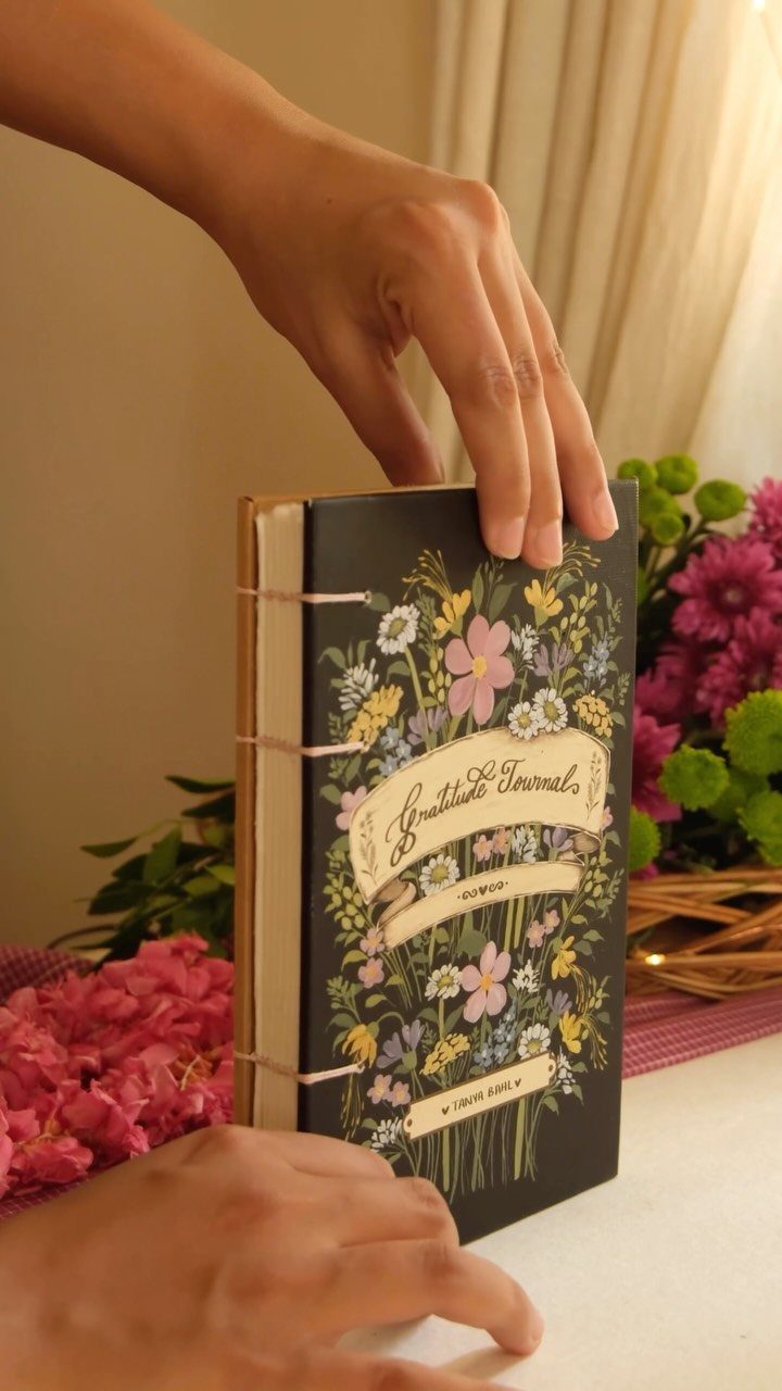 Black gratitude journal with floral cover held by hands near pink flowers