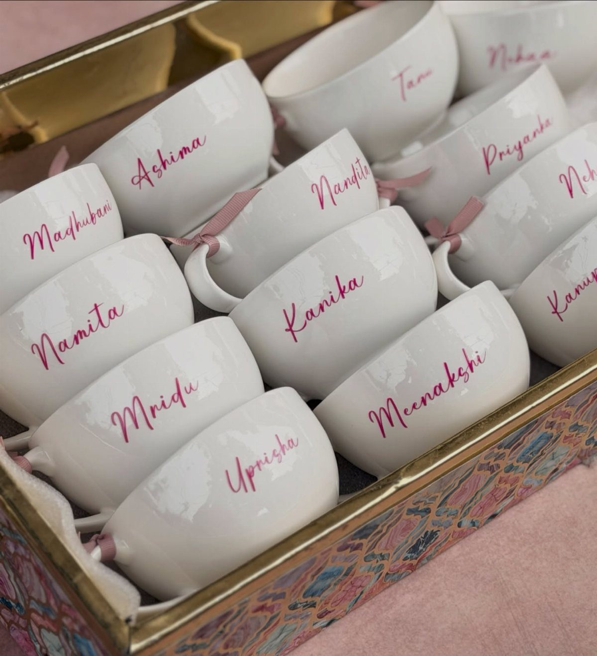 White ceramic mugs with pink personalized names arranged in a decorative gift box.