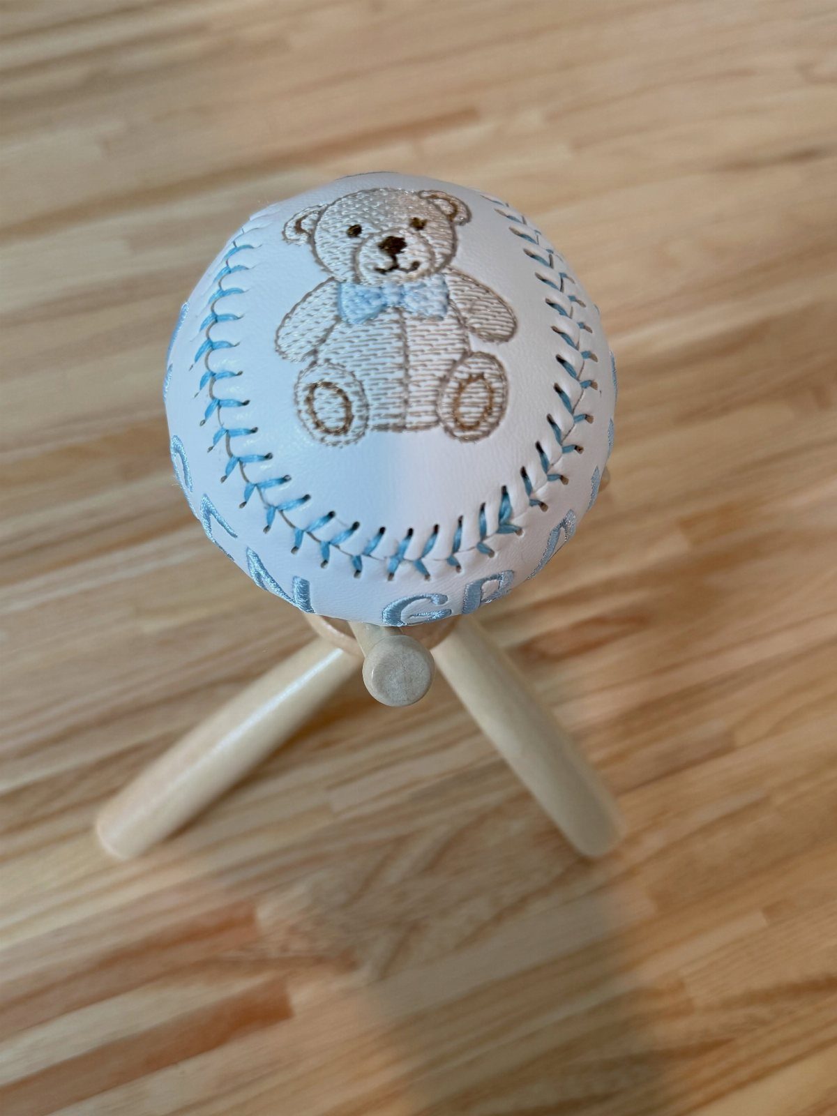 Personalized baseball with an embroidered teddy bear resting on a wooden tripod stand.