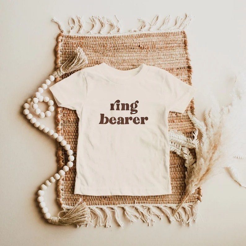 White ring bearer t-shirt on a woven mat with wooden beads and grass.