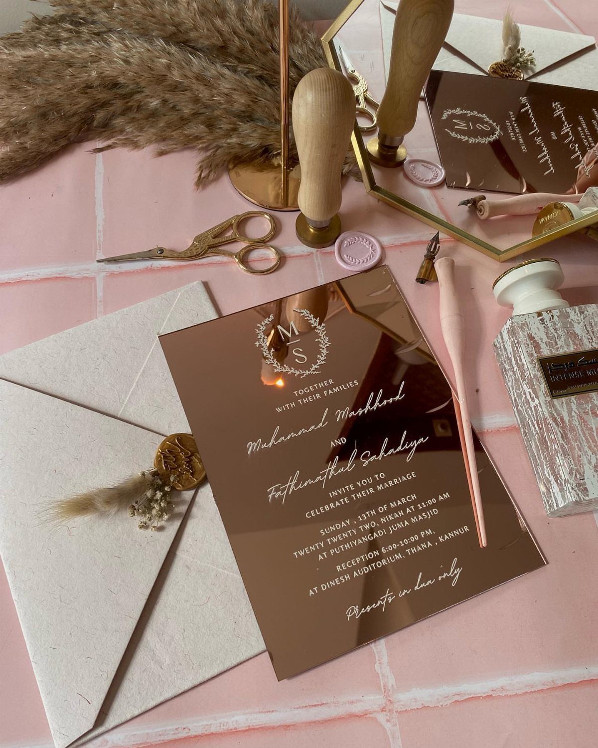 Rose gold mirrored wedding invitation with white text on a pink tiled surface.