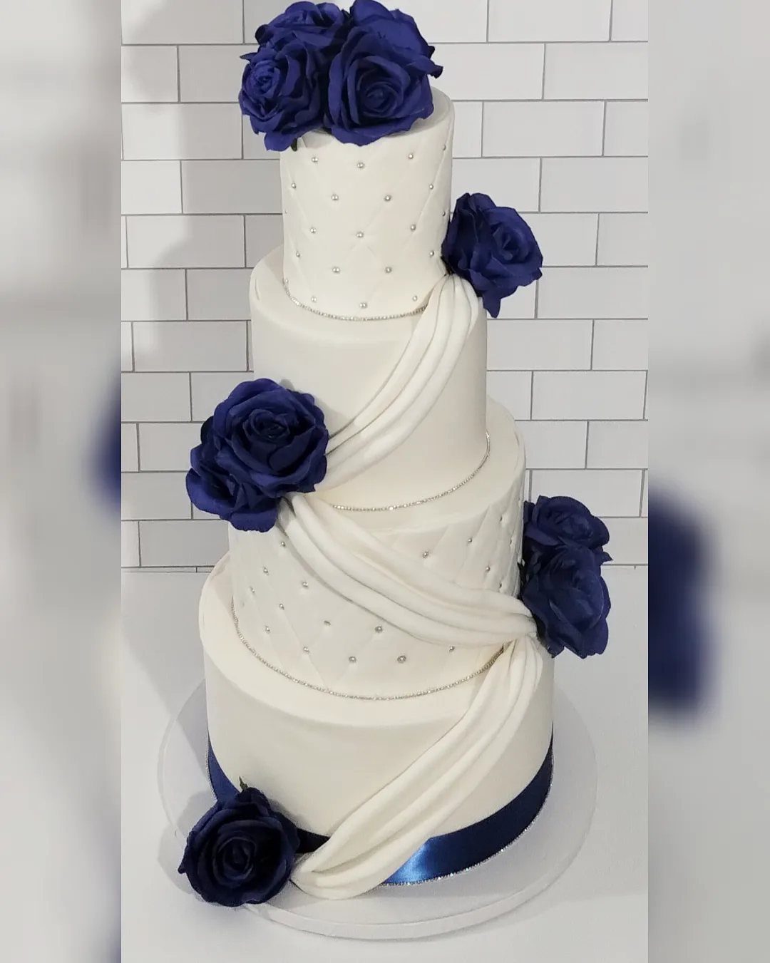 Four-tier white wedding cake adorned with royal blue roses and silver beaded details.