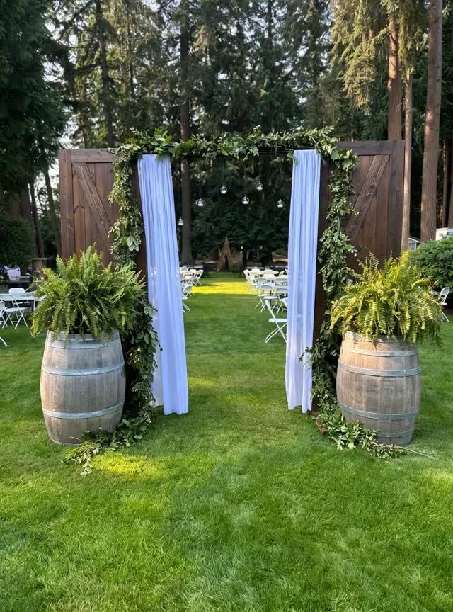 Wooden wedding arch with white fabric and greenery flanked by barrel planters.