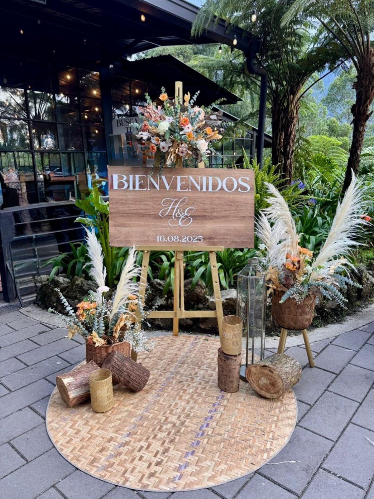 Wooden welcome sign on an easel surrounded by rustic floral arrangements and logs.