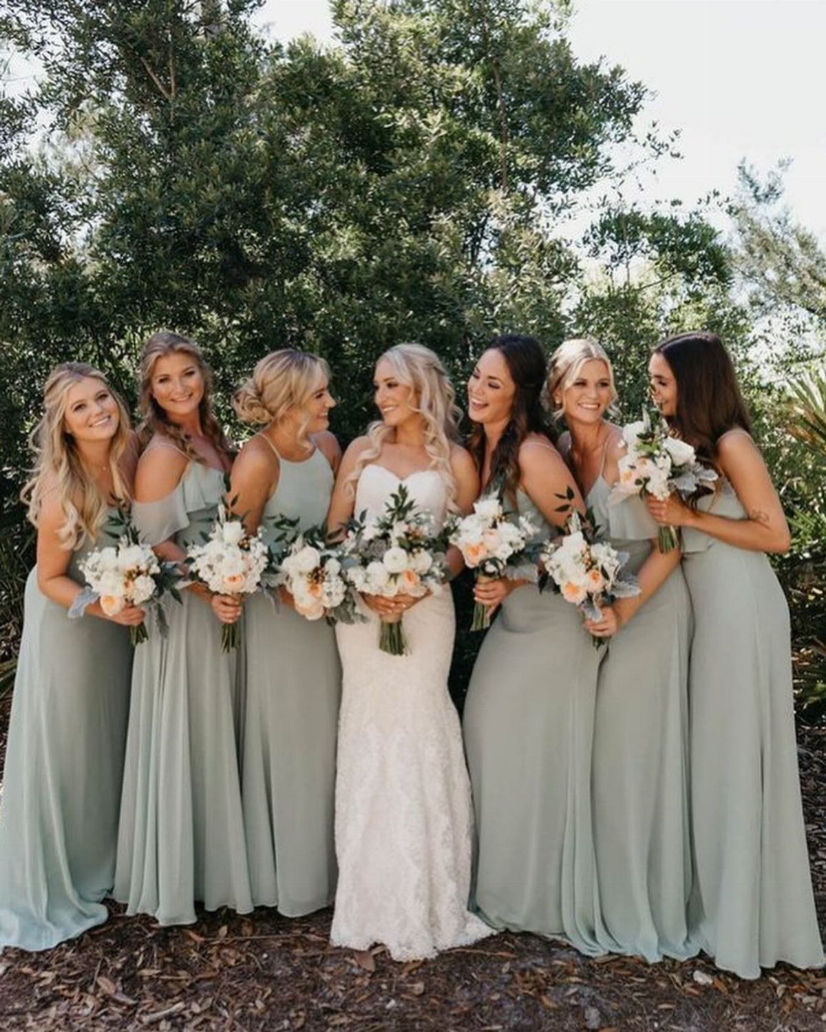 Bride in white lace gown with bridesmaids wearing sage green dresses holding bouquets.