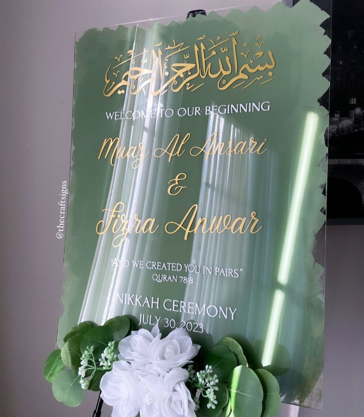 Sage green acrylic welcome sign with gold lettering and white floral accents.