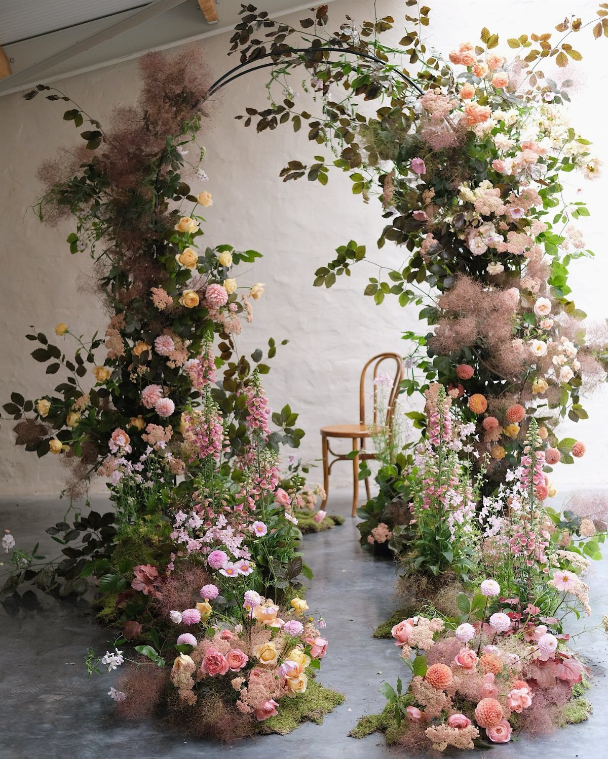 Floral archway with pink and yellow blossoms framing a wooden chair indoors.