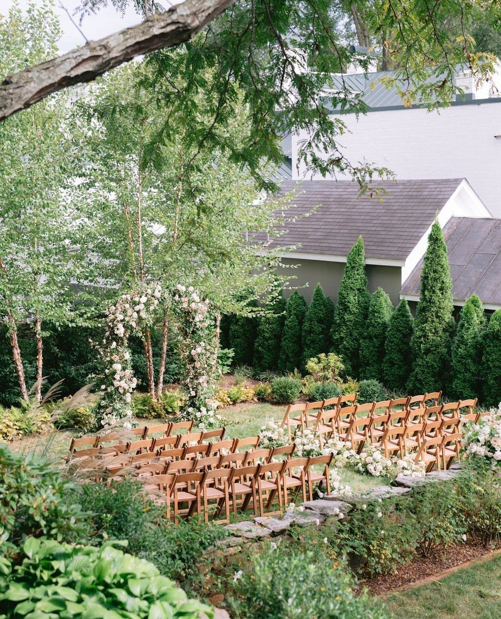 Outdoor wedding ceremony setup with wooden chairs and a white floral archway in a garden