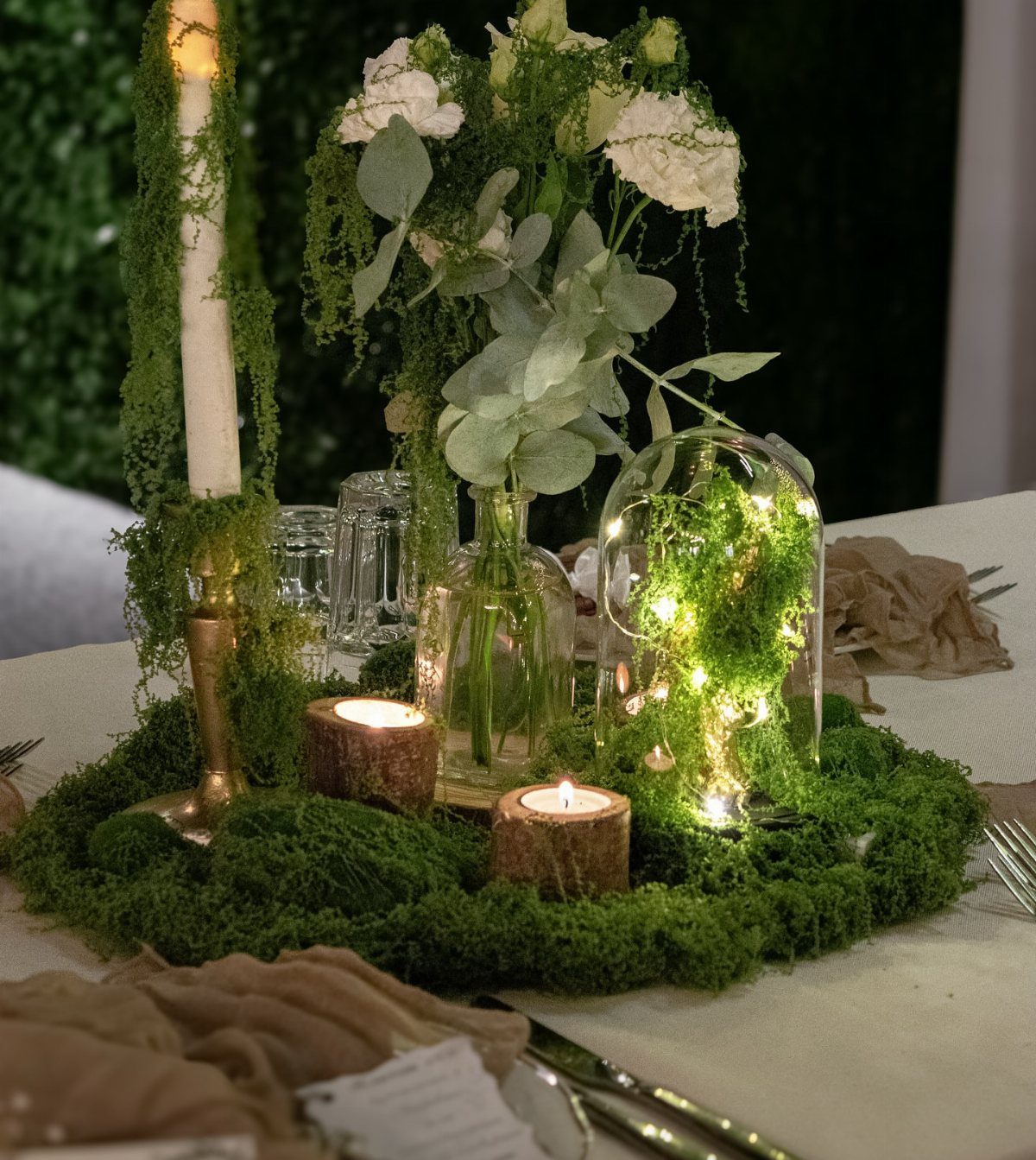 Moss-covered table centerpiece with white flowers, candles, and a glowing glass cloche.