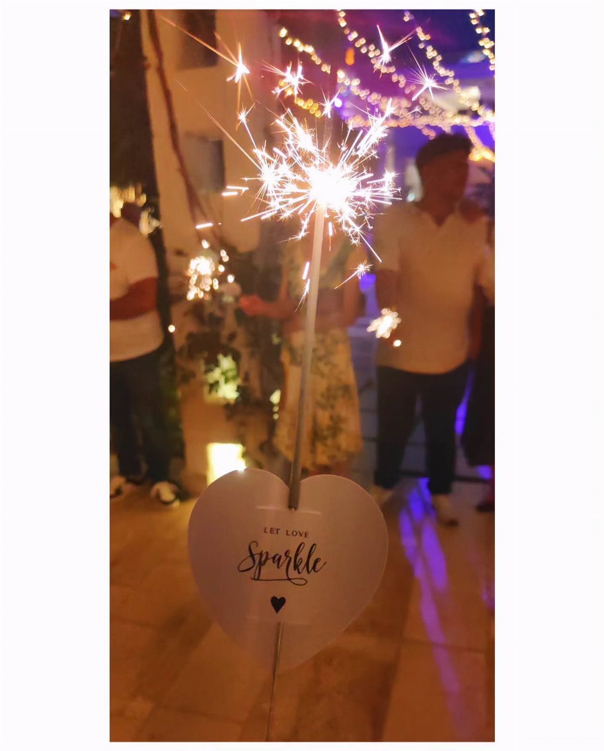 Heart-shaped sign reading Let Love Sparkle attached to a lit wedding sparkler