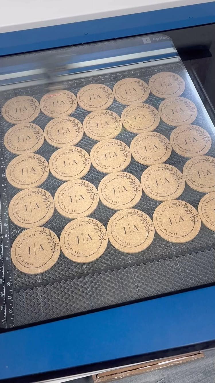 Circular wooden coasters with engraved initials and wreaths inside a laser engraver.