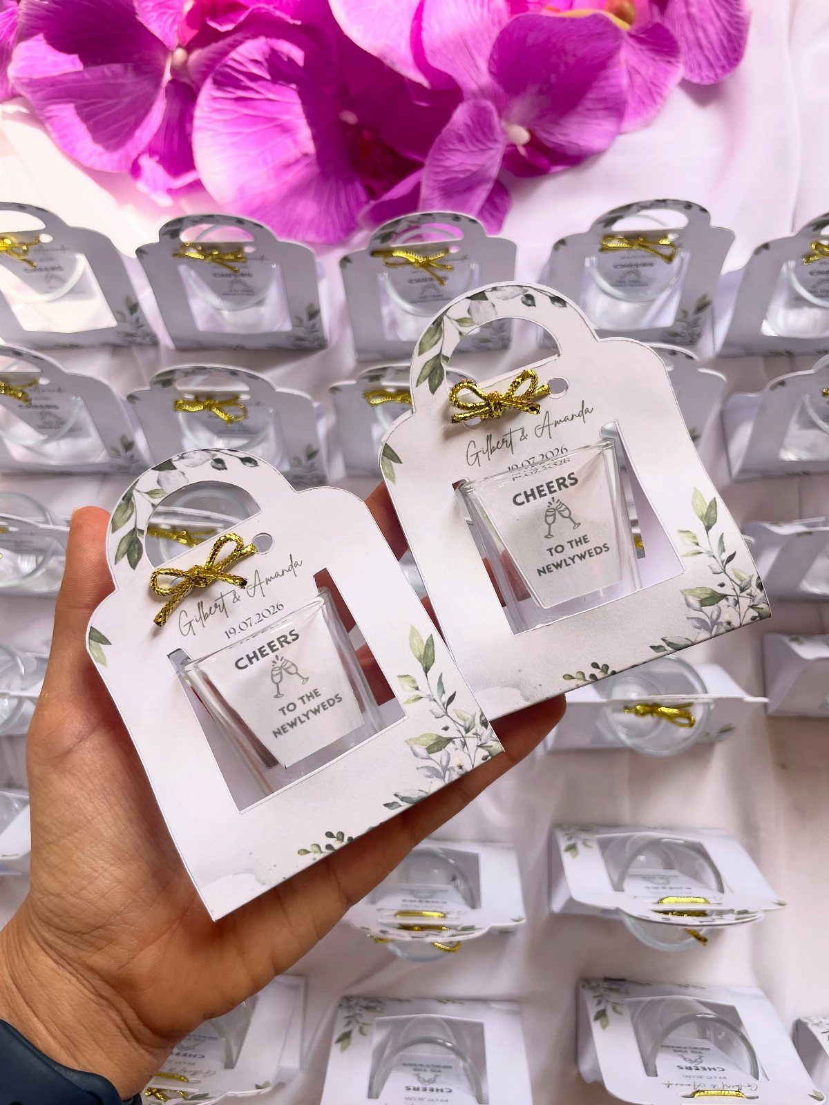 Engraved shot glasses in white decorative holders with gold bows and floral prints.