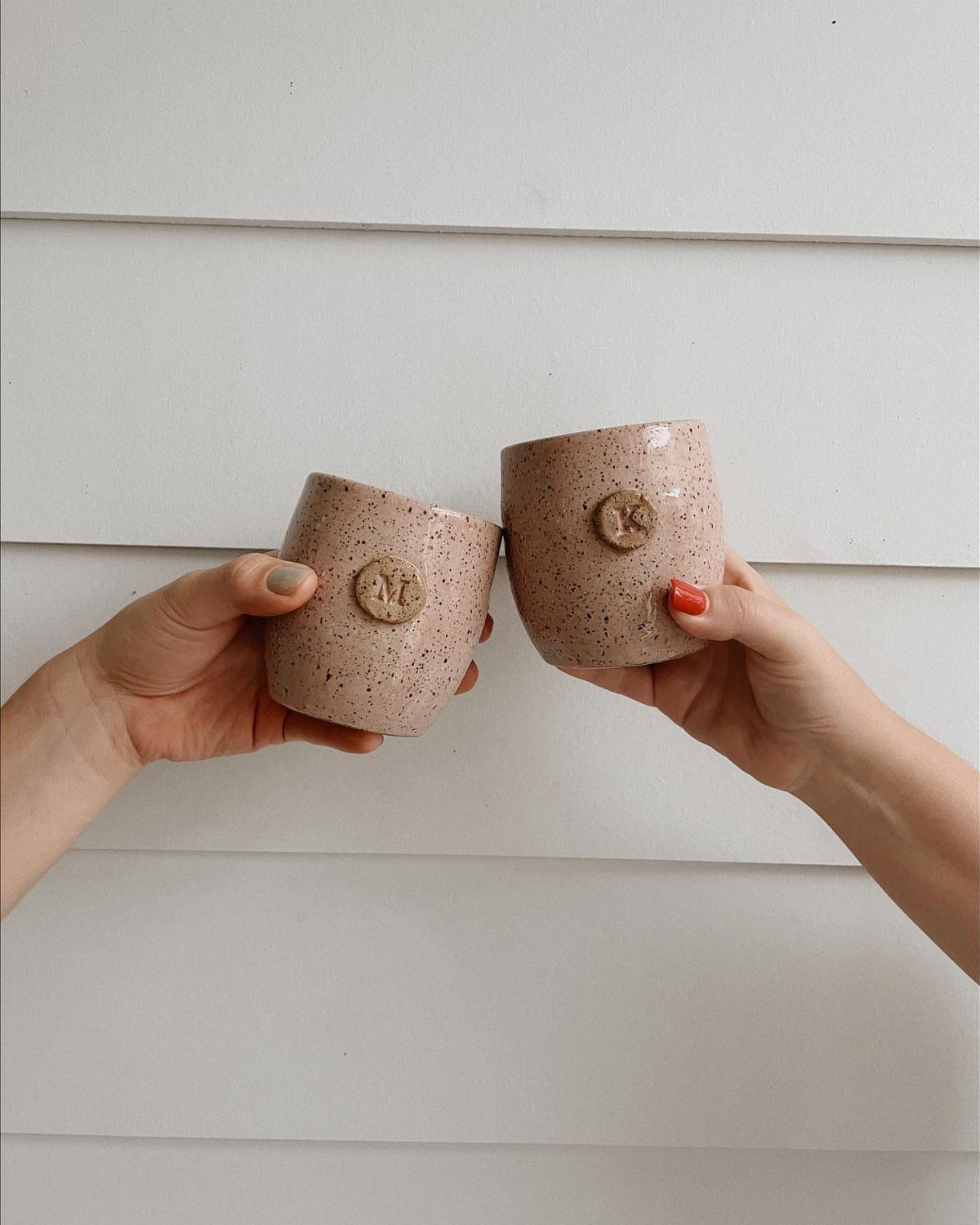Two speckled beige ceramic tumblers with embossed initials held against a white wall.