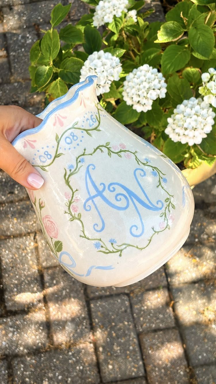 Hand-painted white ceramic pitcher with blue initials and floral designs held outdoors.