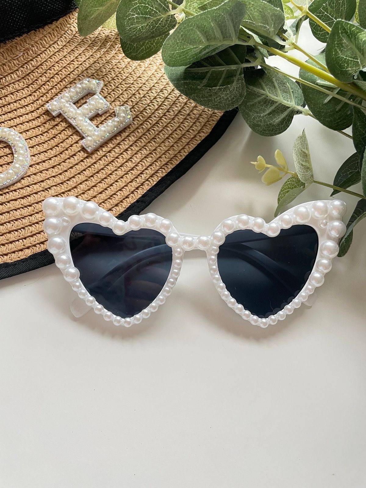 Heart-shaped pearl sunglasses next to a straw hat and green eucalyptus leaves.