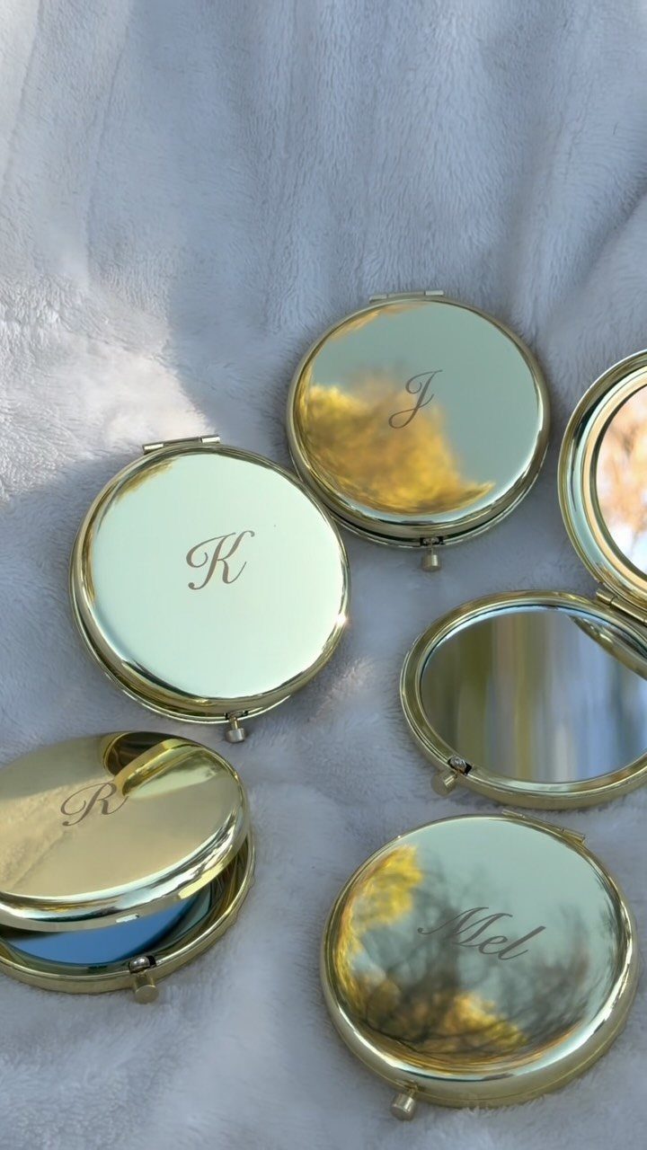 Gold compact mirrors with engraved initials resting on a white fabric background.