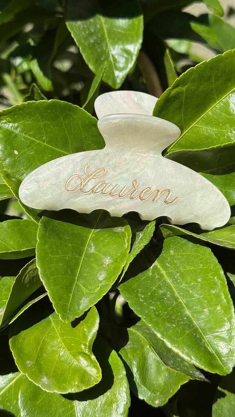 White pearlescent hair claw clip with gold cursive name resting on green leaves