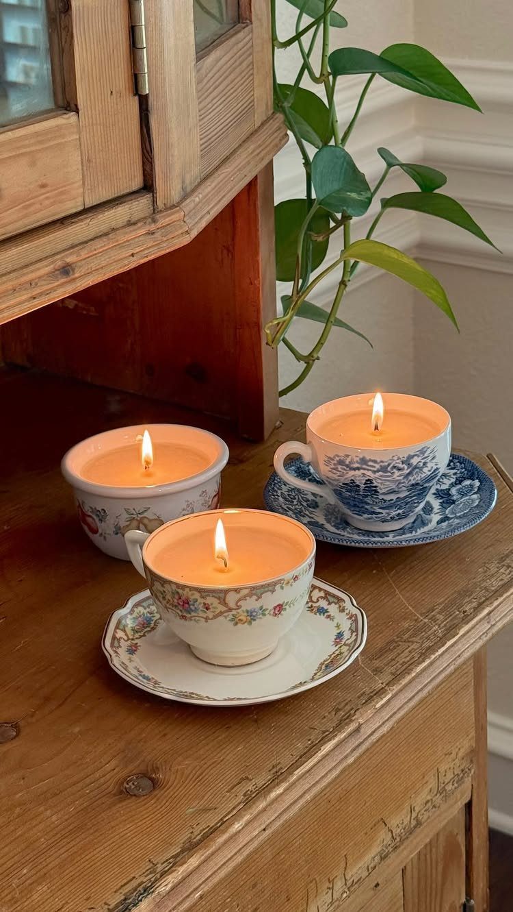 Three lit candles in vintage floral tea cups on a rustic wooden table.