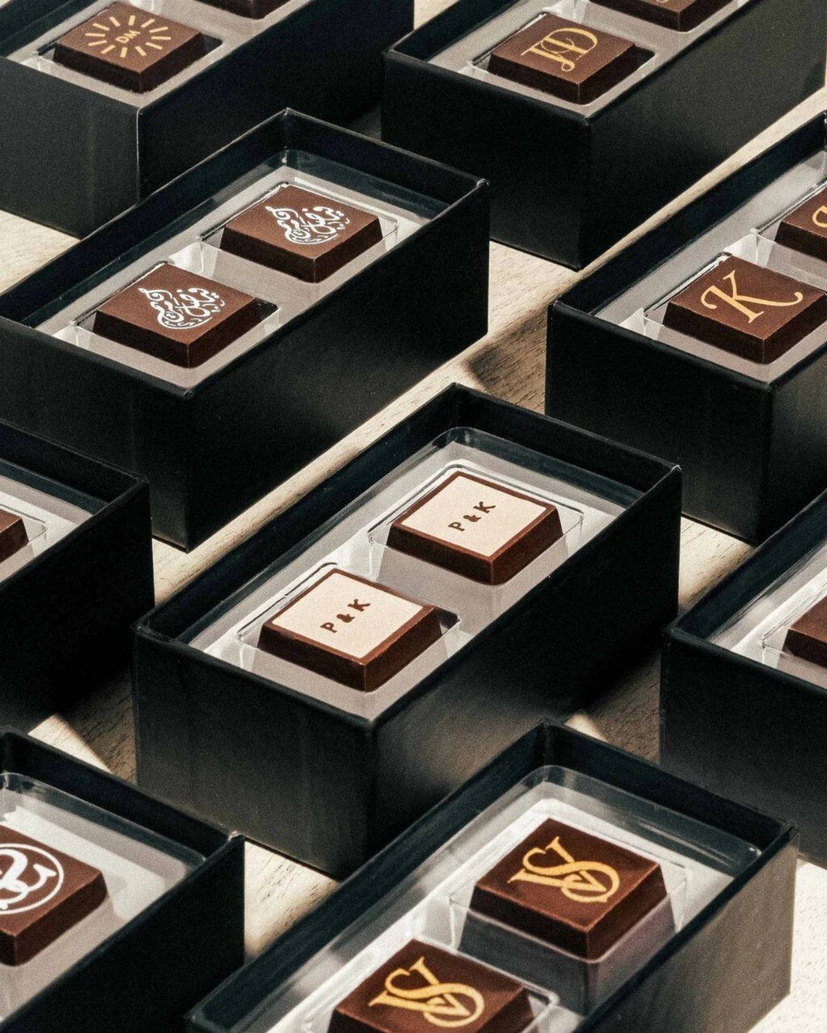 Square brown chocolates with gold initials in small black gift boxes.