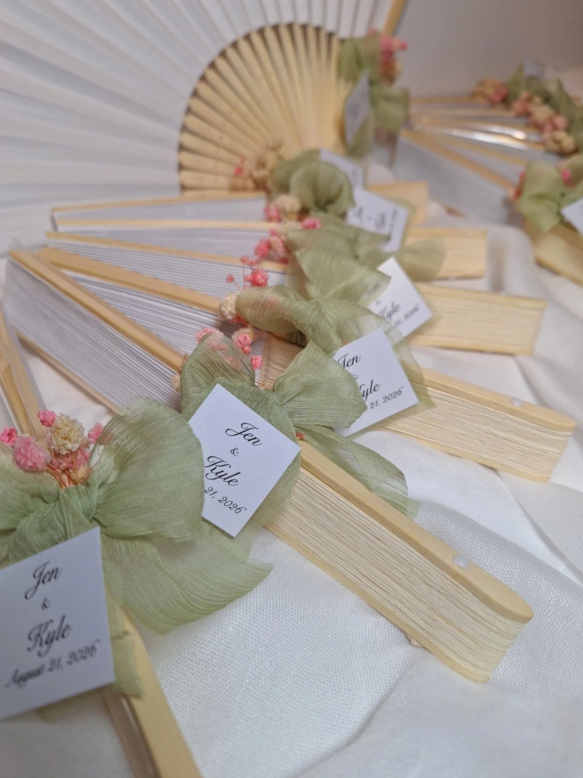 White hand fans with green ribbons and personalized tags for wedding guests.