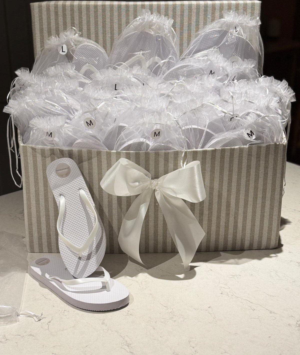 White flip flops in organza bags inside a striped box with a white bow.