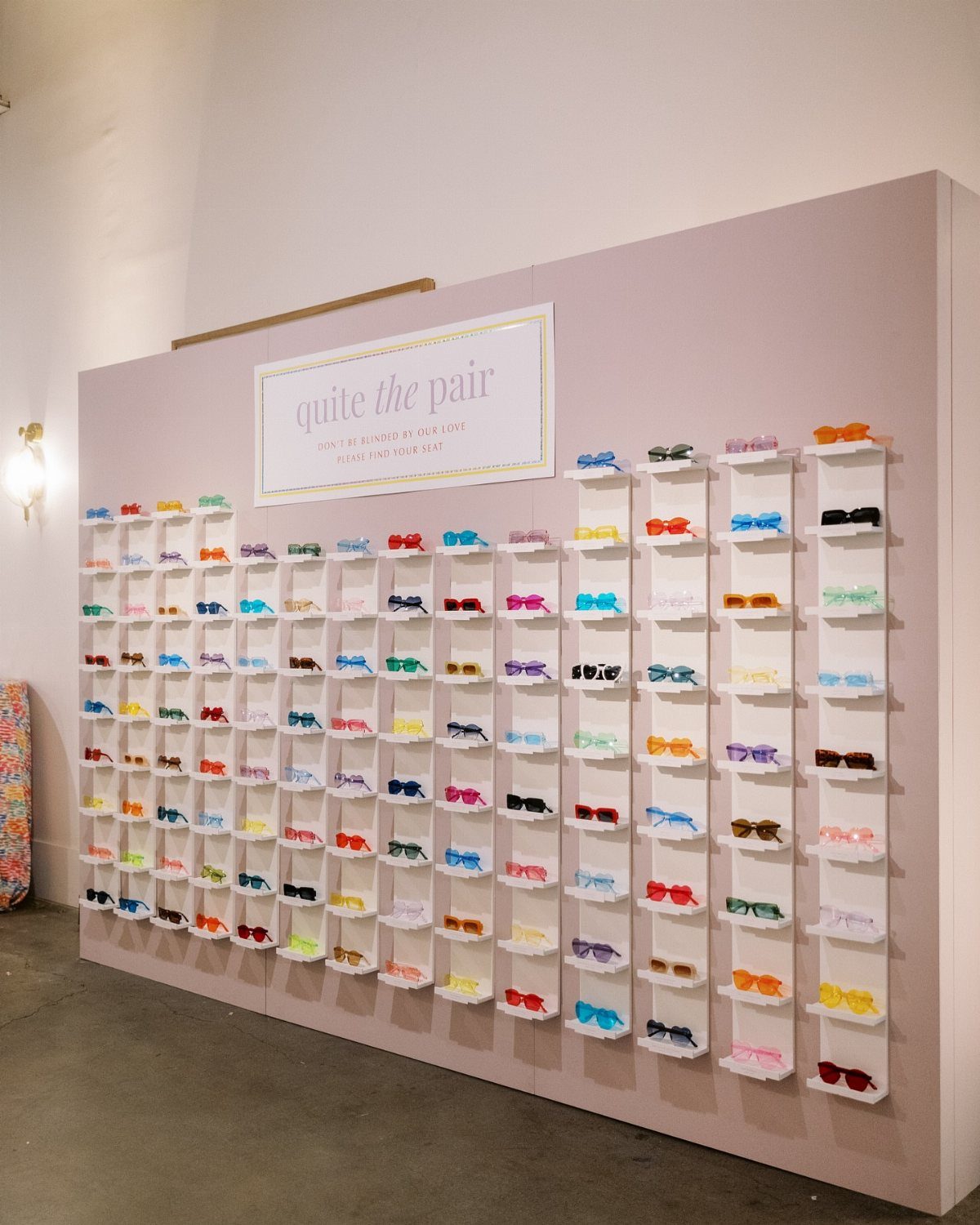 Colorful sunglasses displayed on white shelves against a pink wall with a sign.