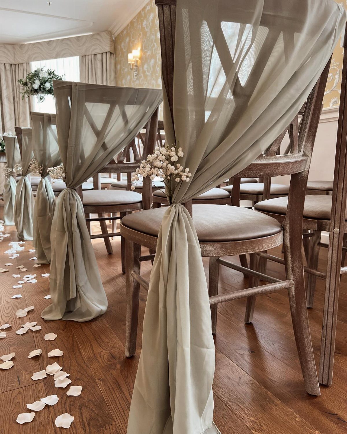Sage green chiffon fabric draped between wooden wedding chairs with scattered white petals.