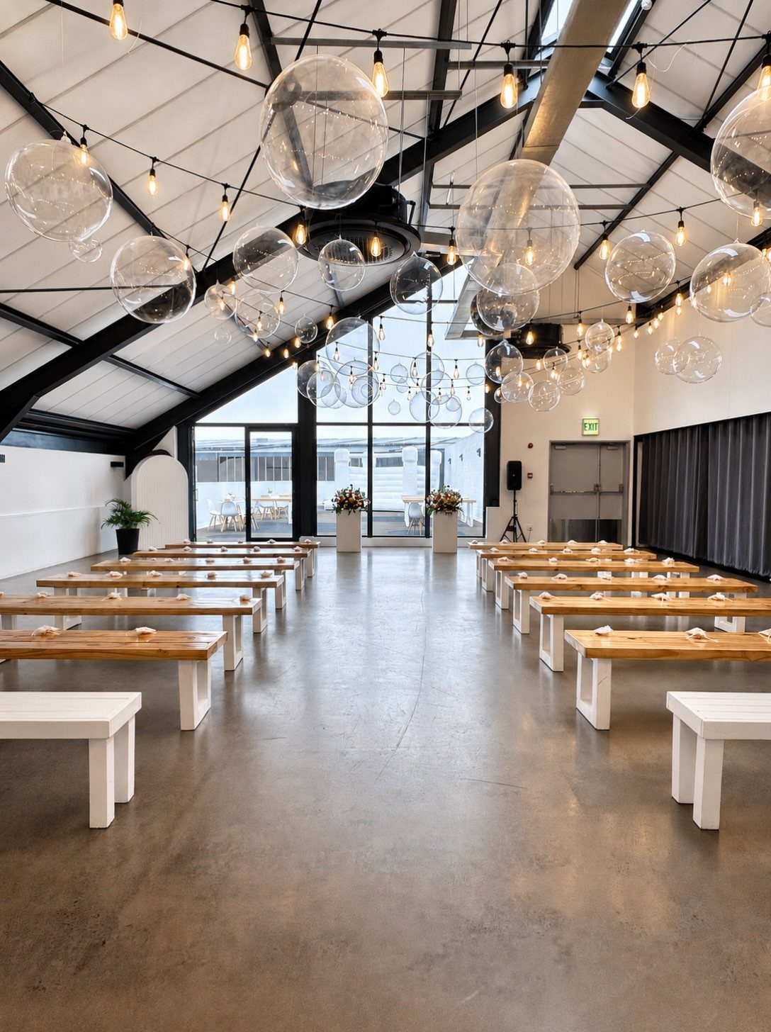 Wooden benches lining a concrete aisle under hanging glass globes and string lights.