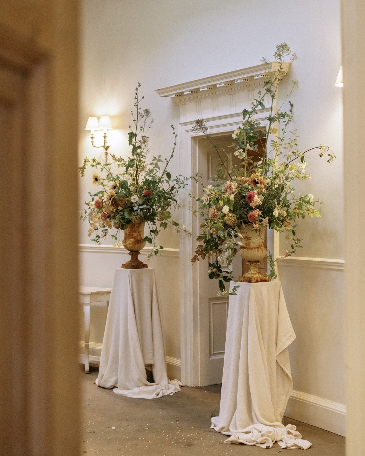 Large gold urns with floral arrangements on white draped pedestals in a hallway.