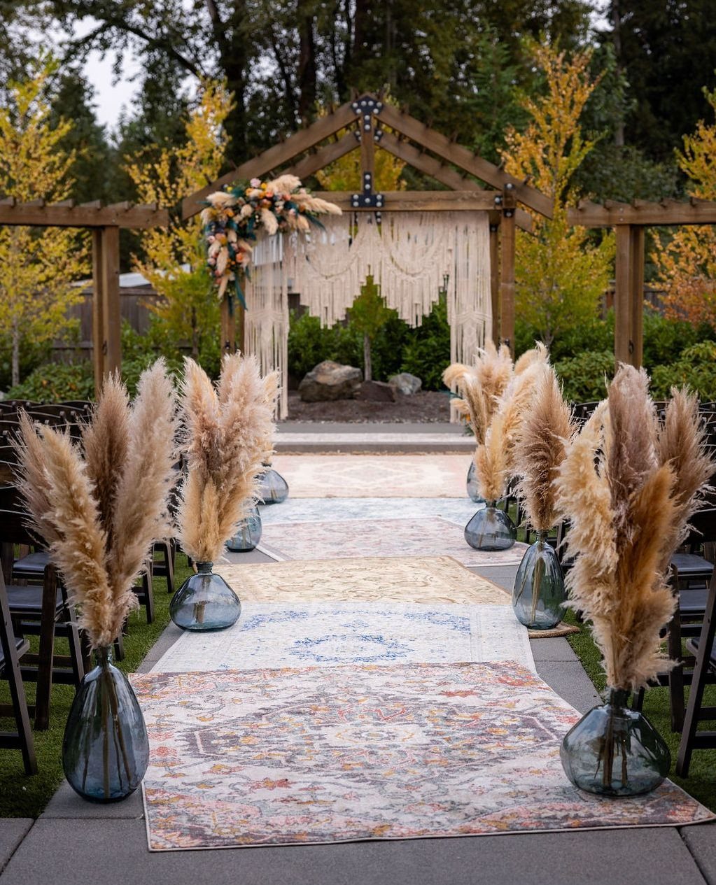 Macrame backdrop on a wooden arbor with pampas grass and patterned aisle rugs.
