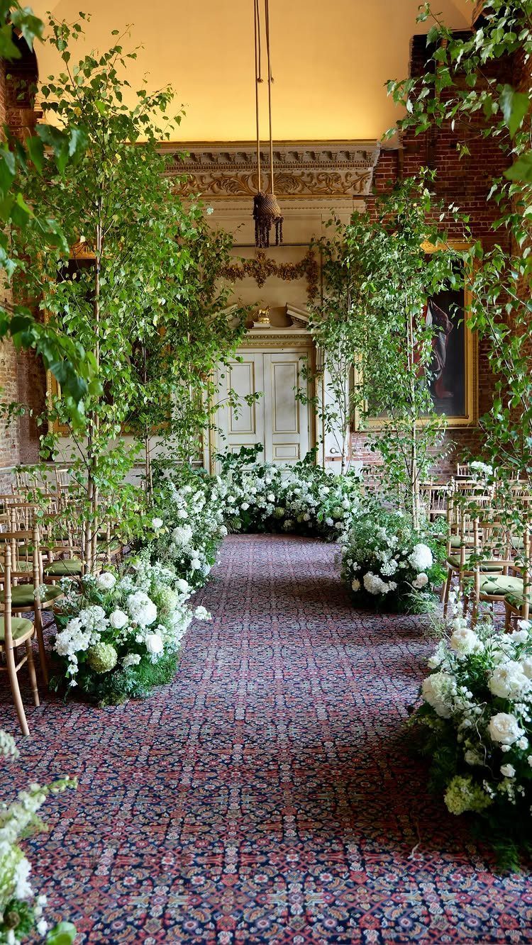 Patterned rug aisle runner lined with white floral arrangements and tall green trees.