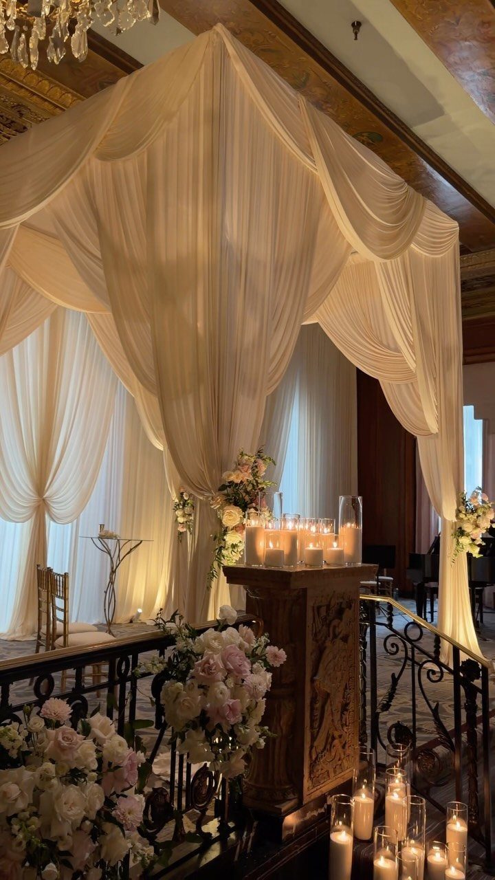 White draped fabric chuppah canopy decorated with pink flowers and glowing candles indoors.