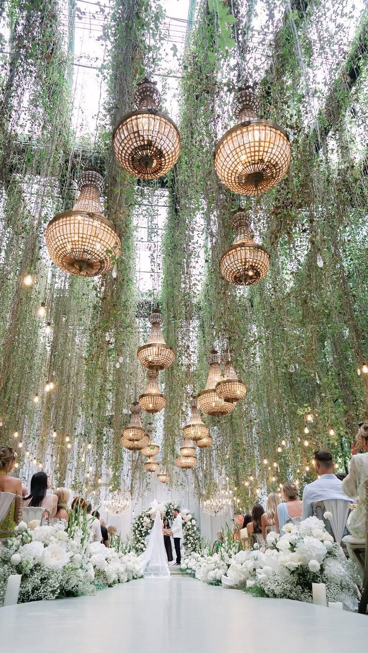 Wedding ceremony aisle with white flowers and hanging greenery and gold chandeliers.