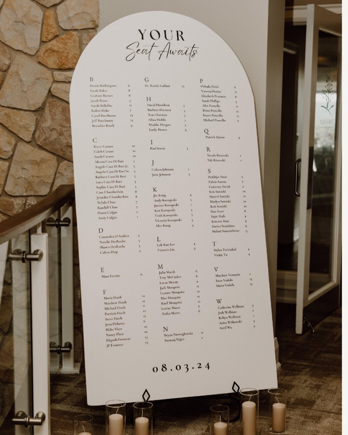 Arched white wedding seating chart sign with guest names and pillar candles below.