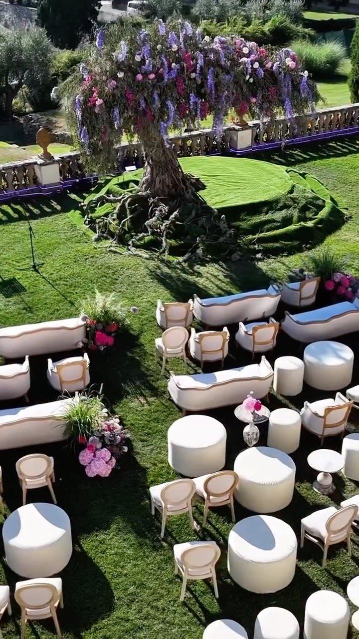 White sofas and ottomans arranged on a green lawn under a flowering tree.
