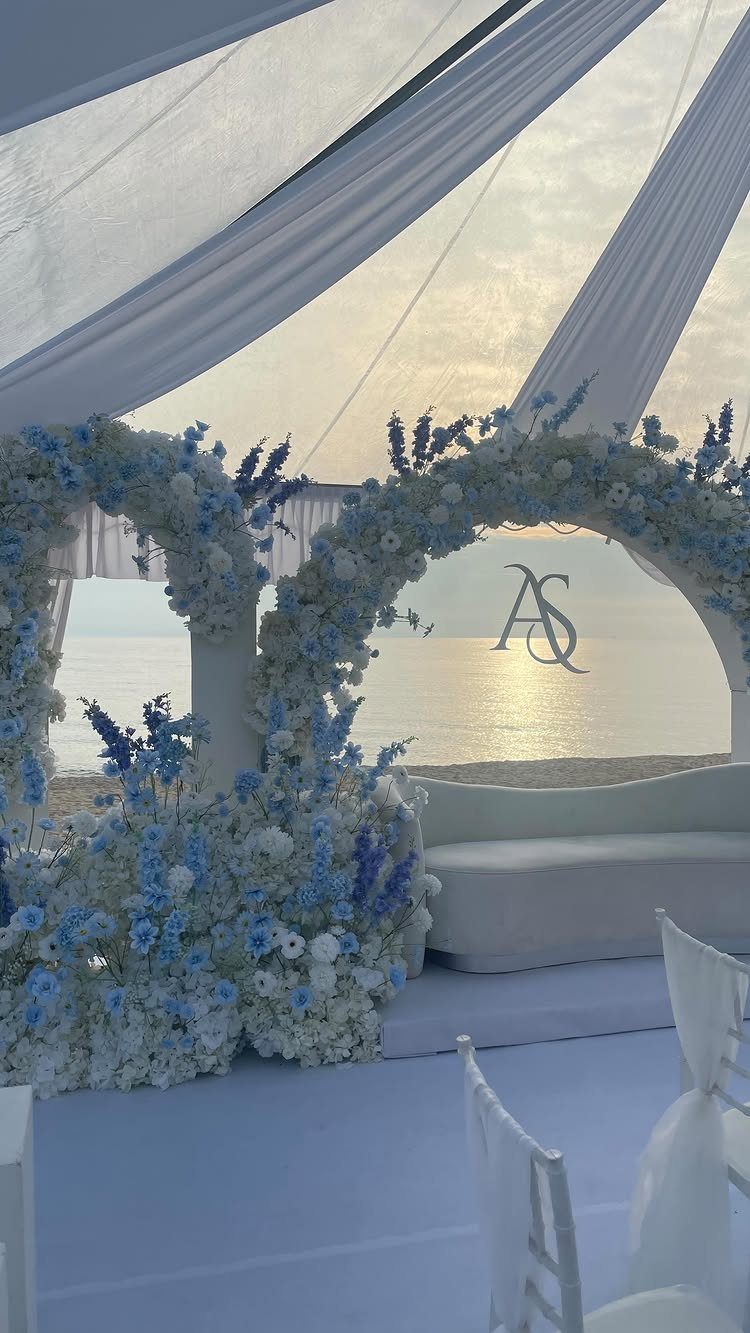 Baby blue and white floral arches under a white canopy on a beach.